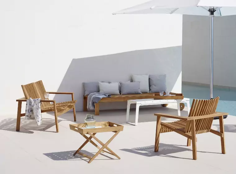 An outdoor sitting area next to a pool with a teak sofa and two teak chairs, a large white umbrella covers the spacee to the right.