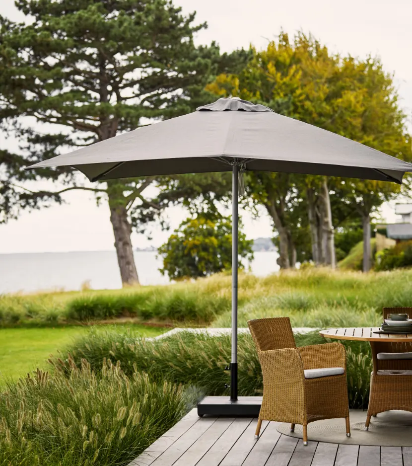 A round teak table with a few chairs on a deck under a large umbrella.