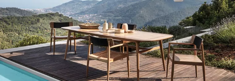 A teak dining table with four chairs on a deck by a pool.