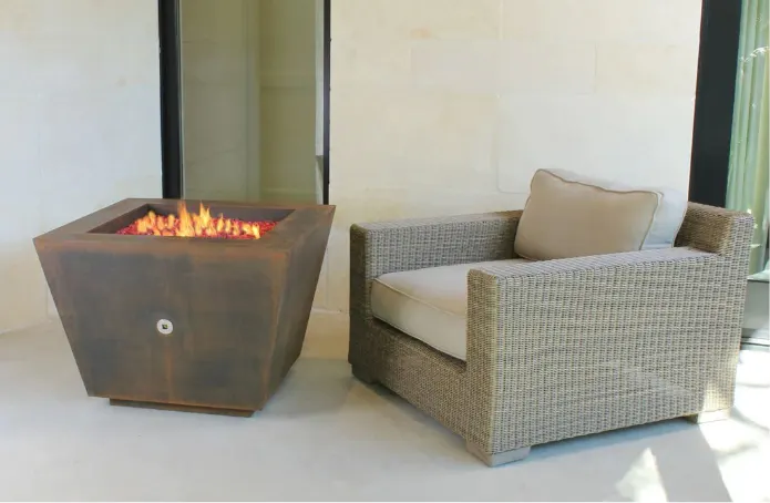 A pyramid shape corten steel fire pit in front of a woven chair.