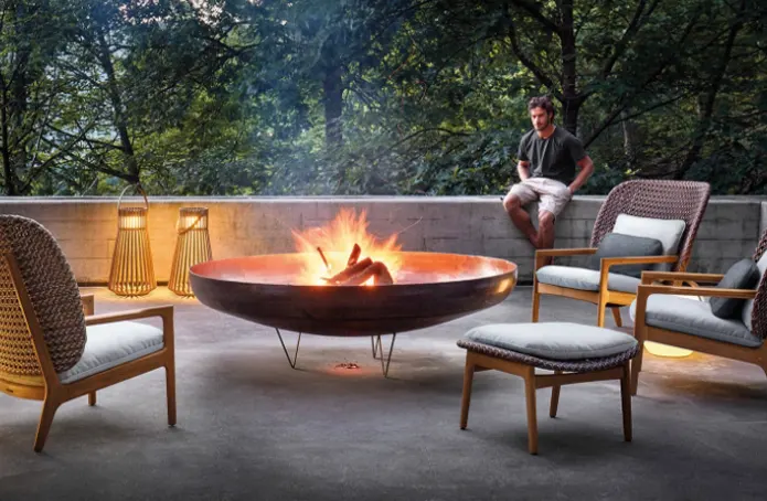 A large fire bowl with burning fire wood while a man sits near it.