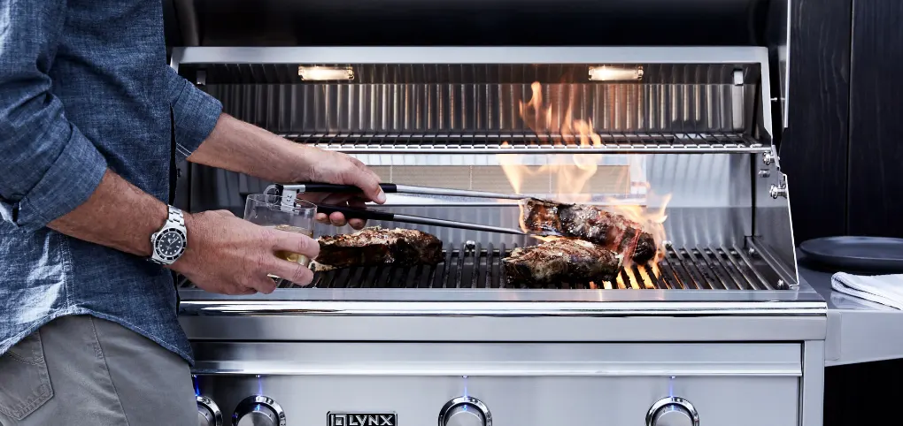 A man grilling steak on a large grill while holding a drink.