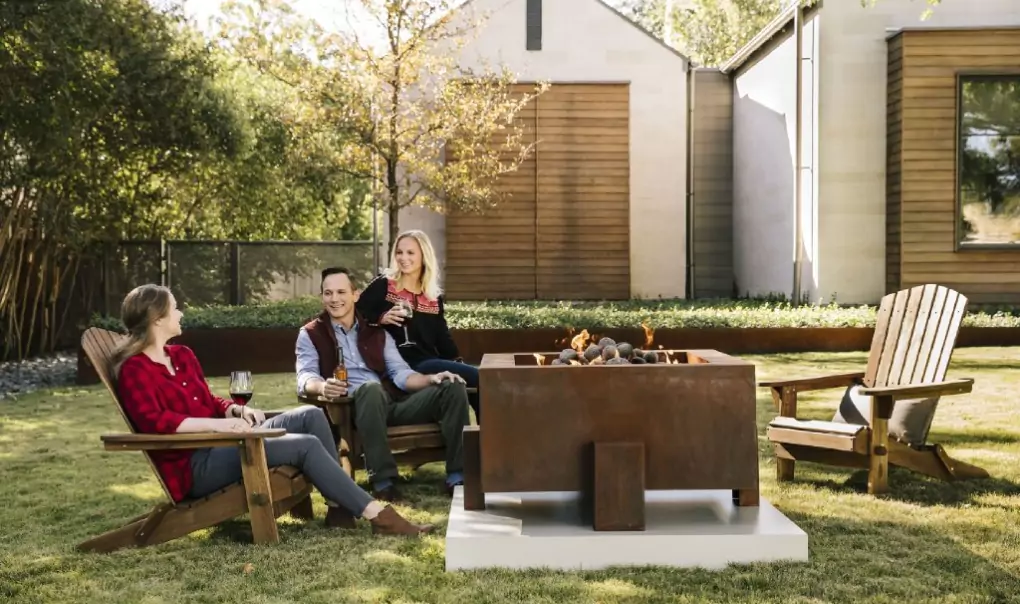 Two woman and a man sitting in front of a fire pit talking.