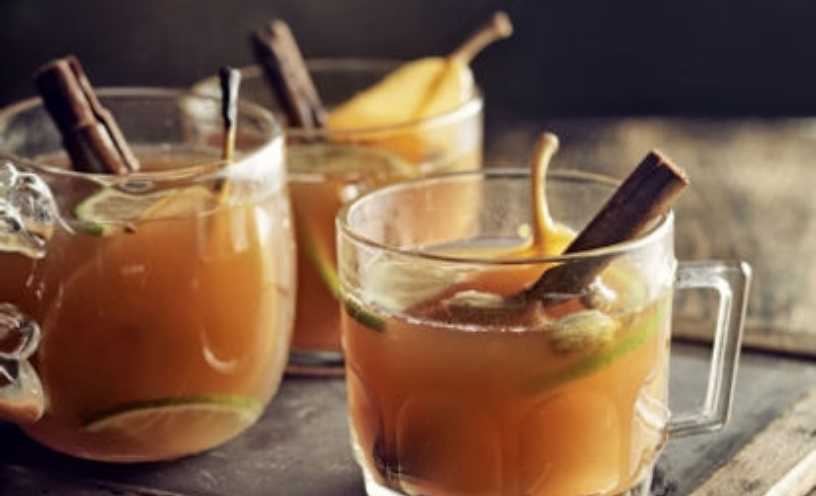 A glass of Apple-Pear Mulled Cider, an orange colored drink with a cinnamon stick and a yellow leaf