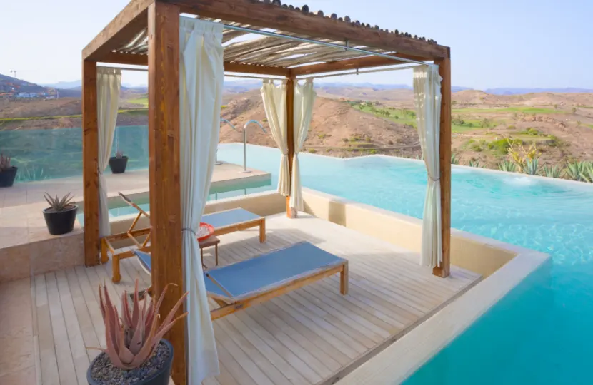 A pergola on a deck with curtains attached to the posts of it and two chaise loungers inside it.