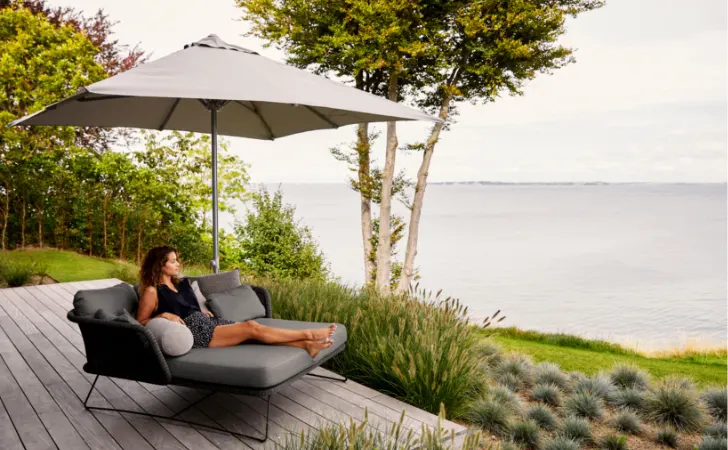 A woman sitting on an outdoor sofa under an umbrella on a deck by the water.