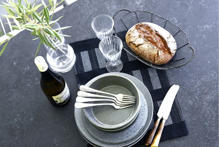 Various plates stacked on top of each other with four forks on them, two glasses, a wine bottle and basket with a baked potato on a table.