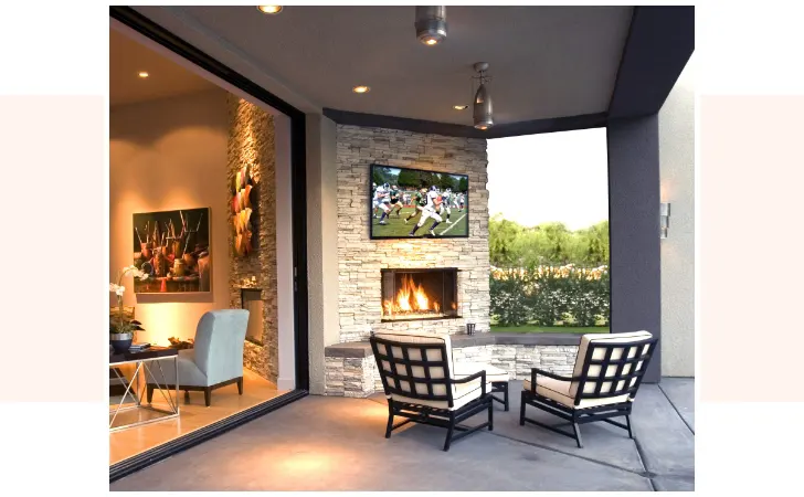 An outdoor sitting area with two chairs in front of a fireplace that has a TV hanging from it.