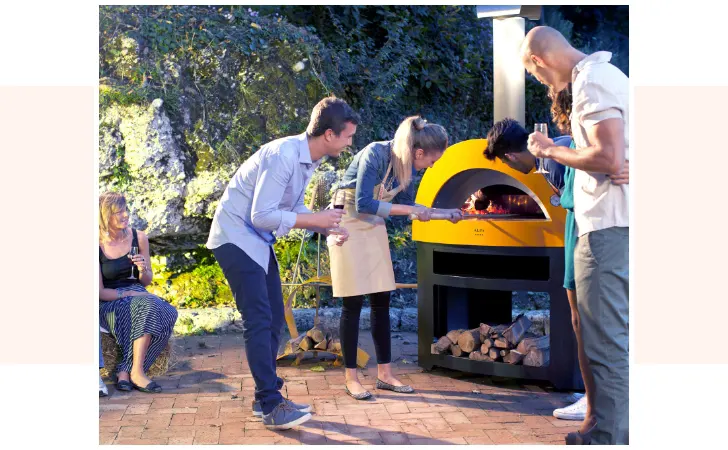 A woman cooking a pizza in a pizza over while various people look at what she's doing.