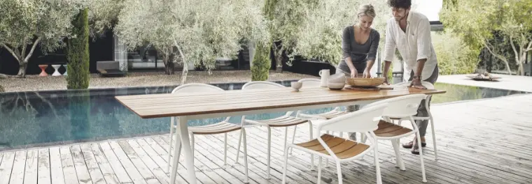 A man and woman standing behind a long rectangular table with six chairs by a pool.
