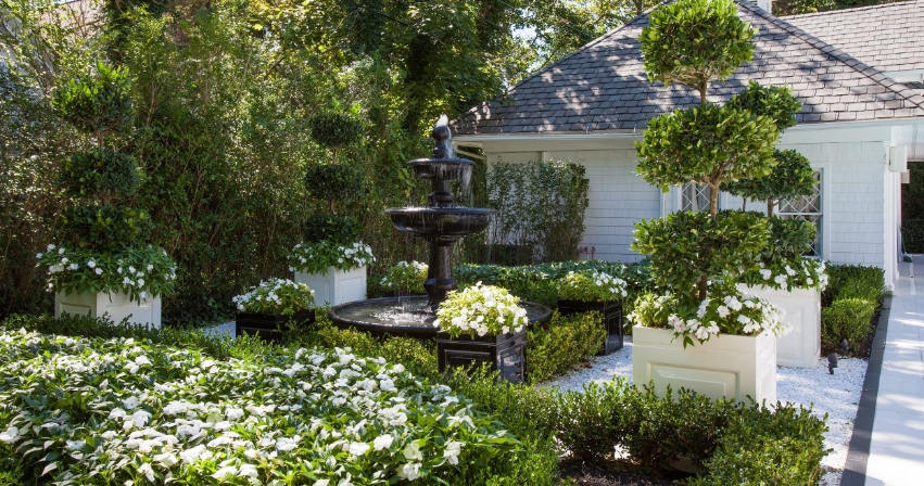 A fountain in the middle of a backyard surrounded by large square planters with trees and various bushes.