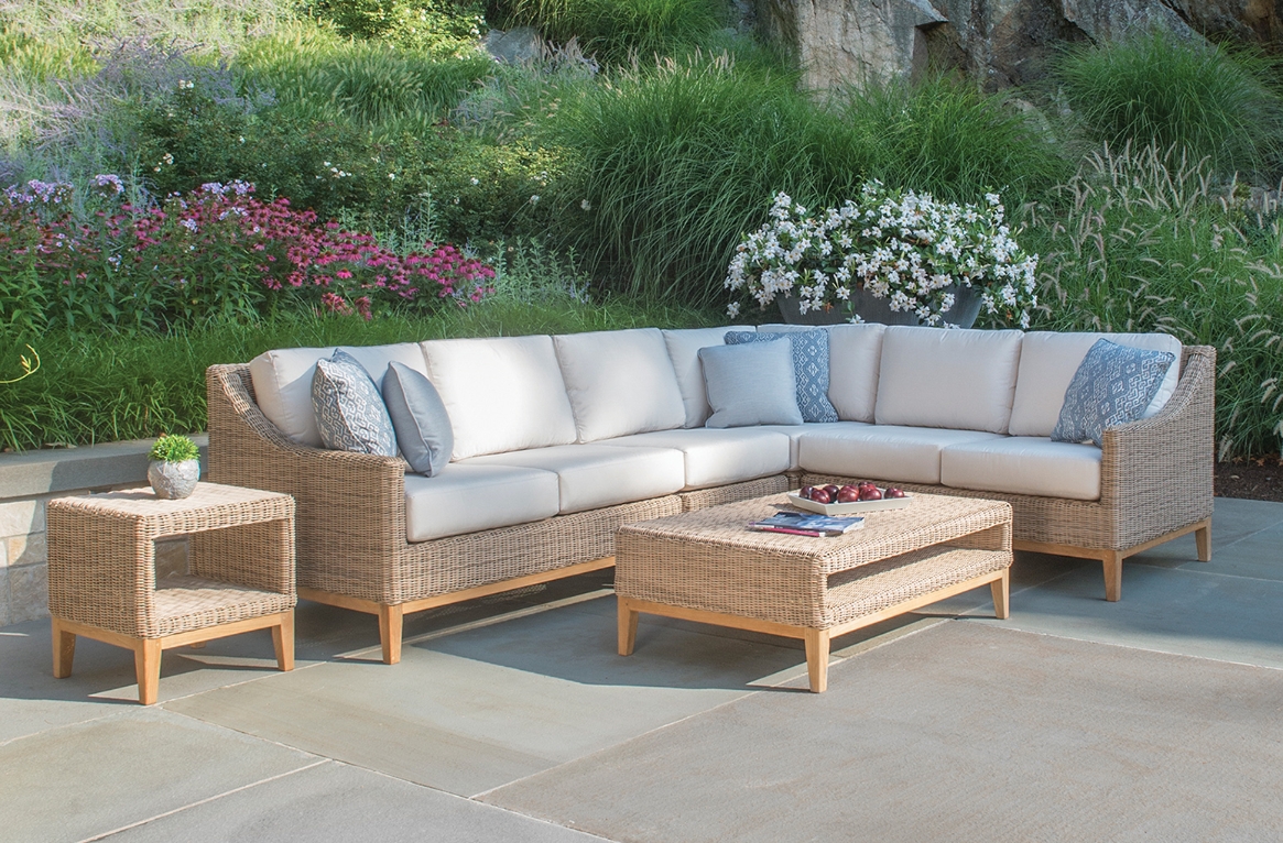 A woven sectional with white cushions with a woven table in front and a small woven side table.
