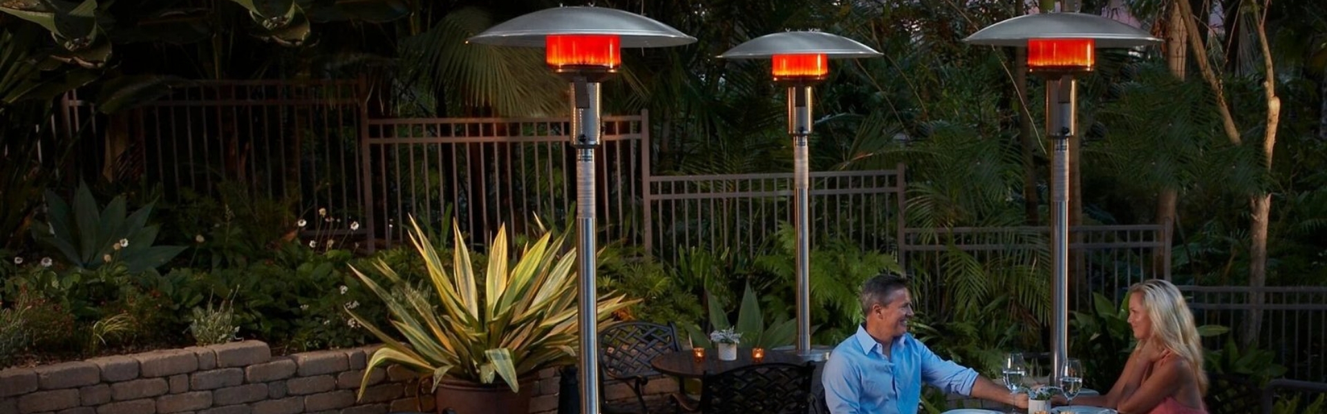 A couple sitting on an outdoor table having drinks by three base standing heaters