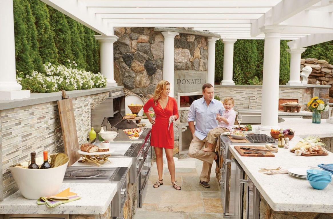 A man and a woman standing in an outdoor kitchen with a little boy sitting on the counter.